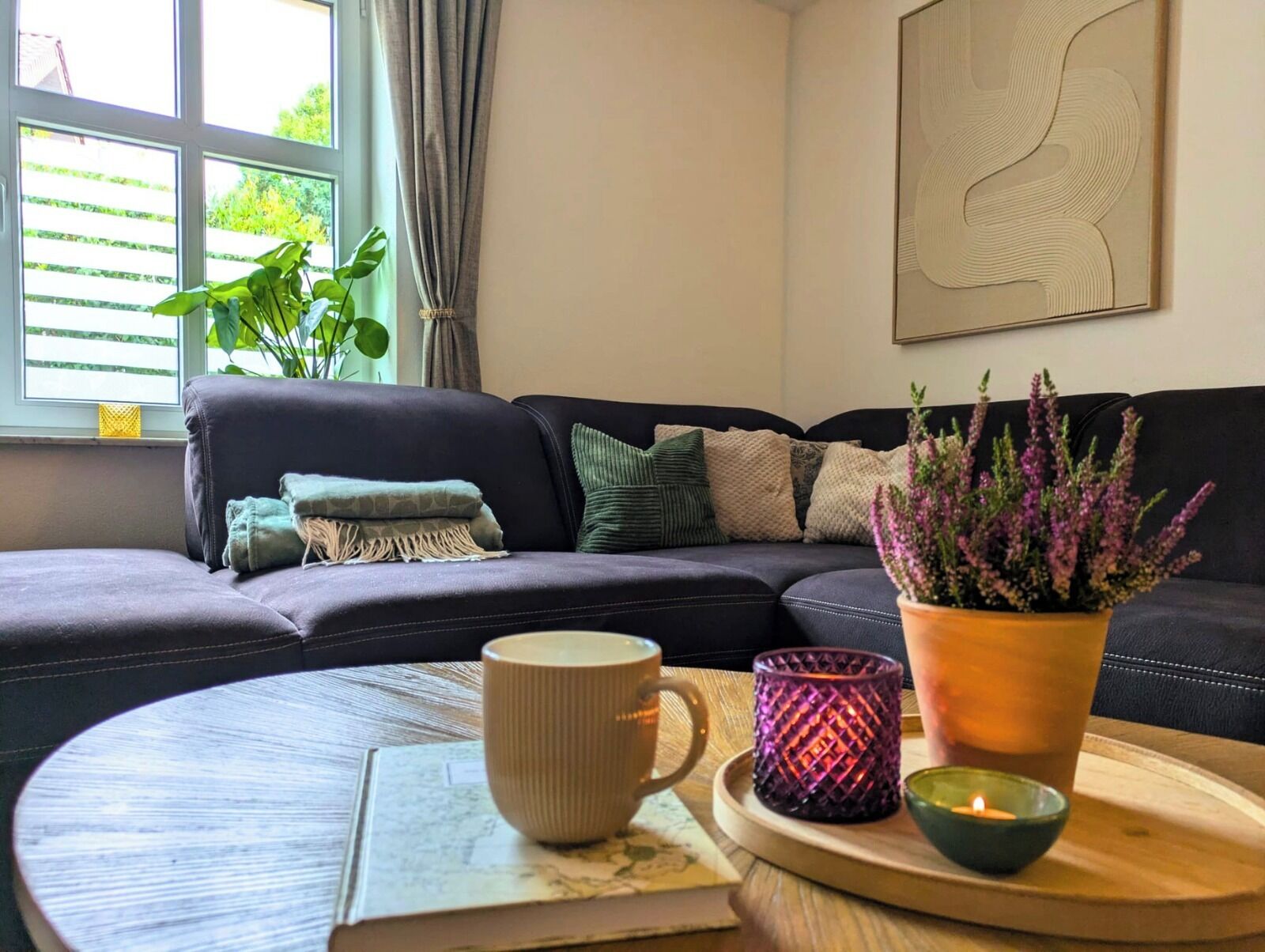 A cozy living room with a dark sofa, a coffee table with a book, a mug, a candle, and a potted plant.