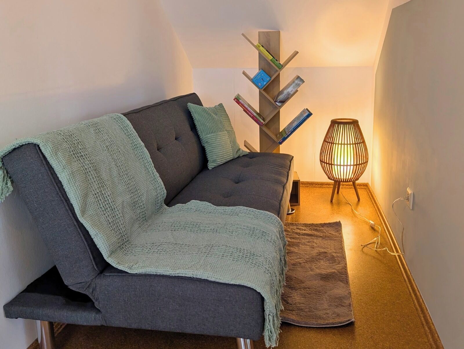 Ett modernt vardagsrum med en soffa, en stickad pläd, ett sidobord med en lampa och en bokhylla.