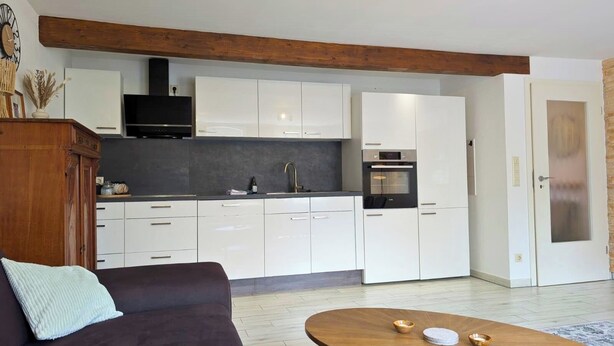 Een moderne keuken met witte kastjes, een houten balk in het plafond, een zwarte oven, en een eethoek met een bruine tafel en twee kommen.