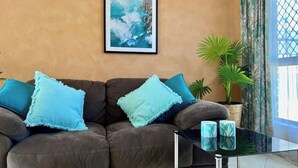 Een zithoek met een bruine bank, twee blauwe kussens, een ingelijste foto aan de muur, een potplant en een glazen salontafel met twee blauwe vazen.