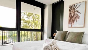 A hotel room with a large bed, a balcony with a view of trees, and a palm tree painting on the wall. - Family apartment in Surfers Paradise (Surfers Paradise)
