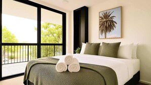 A hotel room with a large bed, a balcony with a view of trees, and a painting of a palm tree on the wall. - Family apartment in Surfers Paradise (Surfers Paradise)