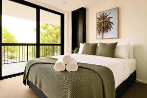 A hotel room with a large bed, a balcony with a view of trees, and a painting of a palm tree on the wall. - Family apartment in Surfers Paradise (Surfers Paradise)