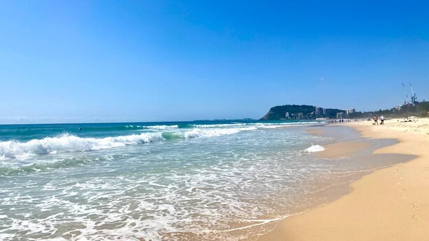 Una playa con olas, cielo despejado y edificios al fondo.