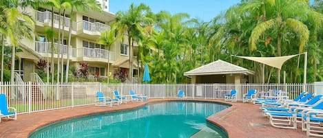 Un área de piscina con sillas reclinables azules, una piscina de ladrillo y un edificio blanco con balcones.