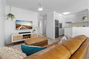 A modern living room with a leather sofa, a flat-screen TV, and a kitchen area with white cabinets and a stainless steel refrigerator. - Beach Escape with Pool & BBQ (Surfers Paradise)