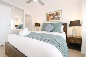 A neatly made bed with a white comforter and a teal throw blanket, two bedside lamps, a framed abstract artwork above the bed, and a wooden nightstand with a drawer. - Beach Escape with Pool & BBQ (Surfers Paradise)