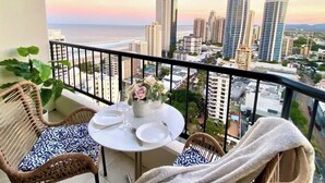 A balcony with wicker furniture, a table set for two, and a view of a cityscape with tall buildings and a sunset. - Top level Ocean View Oasis: 3-Bed&Free Wi-Fi (Surfers Paradise)