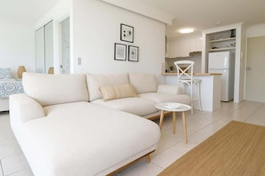 A modern living room with a sofa, a small table, and a kitchen area with cabinets and a refrigerator. - Ocean View 1BR Steps to Beach (Surfers Paradise)