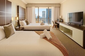 Ein Hotelzimmer mit zwei Betten, einem Fernseher, einem Schreibtisch und Blick auf hohe Gebäude.