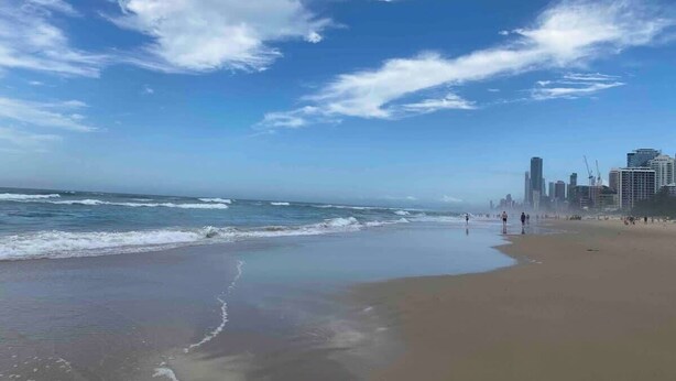Una spiaggia con sullo sfondo una linea di edifici e il cielo sereno, increspata dalle onde.