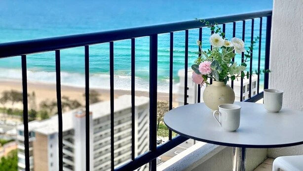 Een balkon met uitzicht op de oceaan en het strand, een vaas met bloemen en twee witte kopjes op een klein tafeltje.