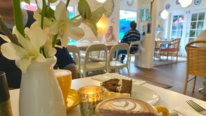 Een eettafel met een gele koffiekop, een plakje taart en een witte vaas met bloemen.
