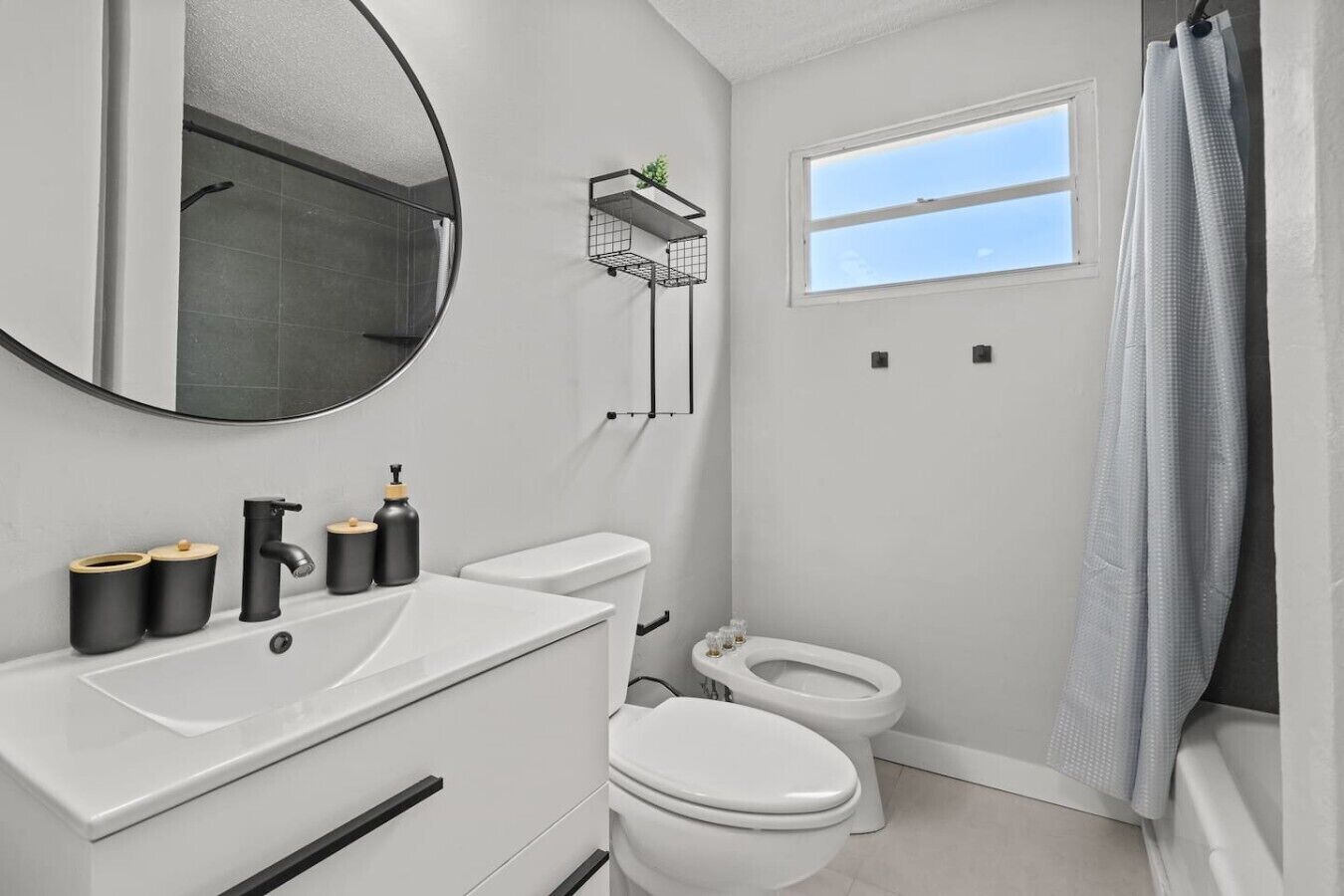 A bathroom with a white sink, a toilet, a shower with a curtain, a window, and a mirror.