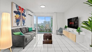 A modern living room with a grey sofa, a coffee table, a TV, and a balcony with a view of the city.