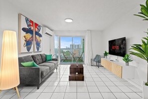 A modern living room with a grey sofa, a coffee table, a TV, and a balcony with a view of the city. - High-Rise Condado Suite | Ocean Views & PR Skyline (San Juan)