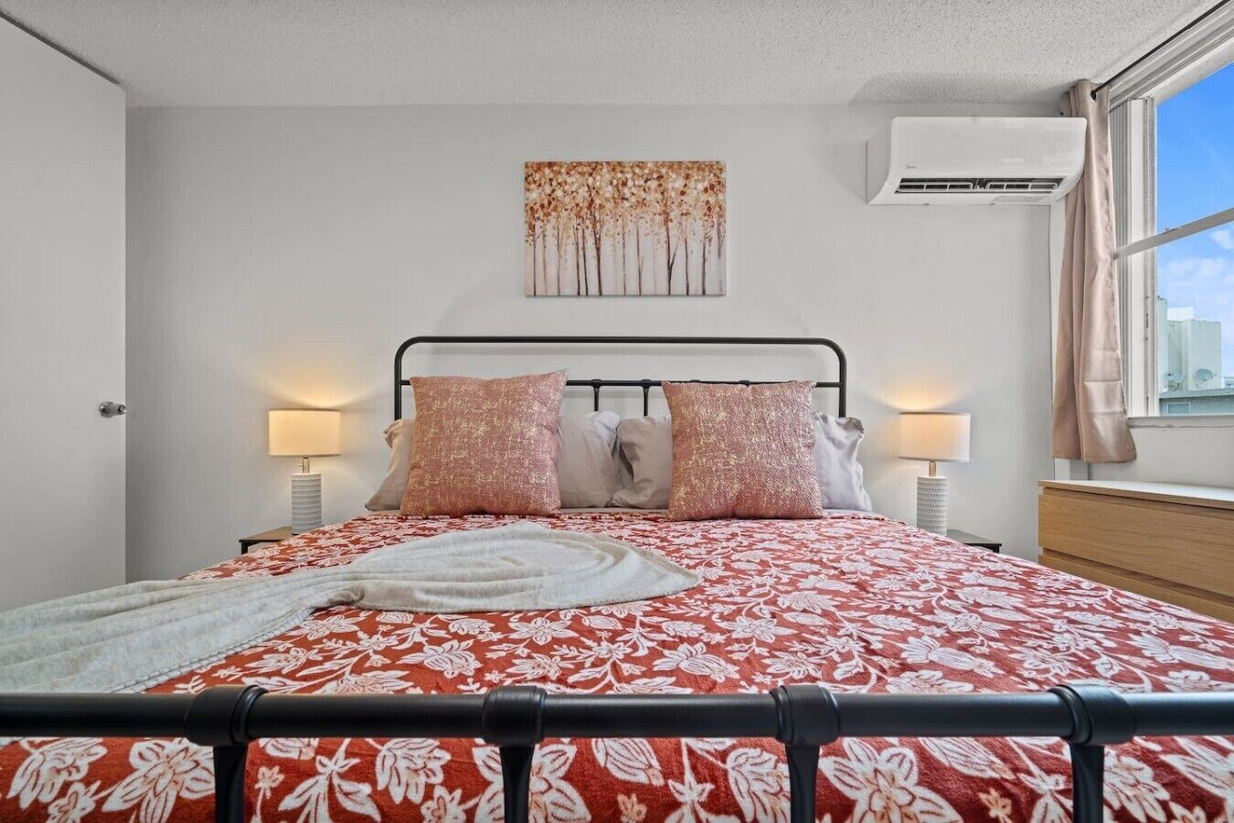 Un lit avec une couvre-lit à motifs floraux, deux lampes de chevet, un climatiseur fixé au mur et une fenêtre avec des rideaux.
