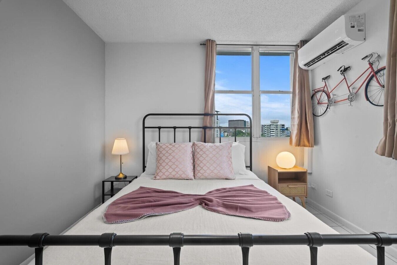 Una camera da letto con un letto, due comodini, una finestra con tende, un condizionatore e una bicicletta appesa al muro.