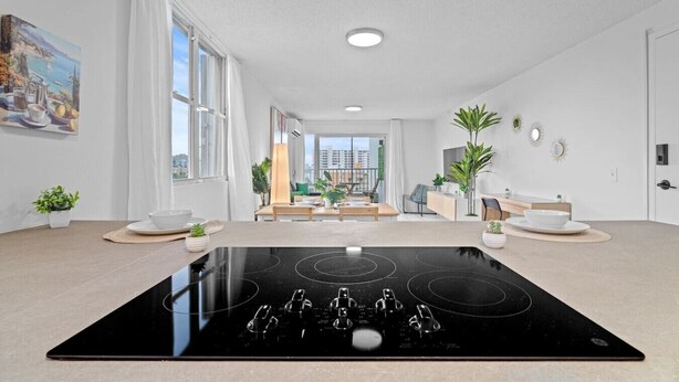 A modern living room with a black induction cooktop, large windows, a dining area, and a view of the cityscape.