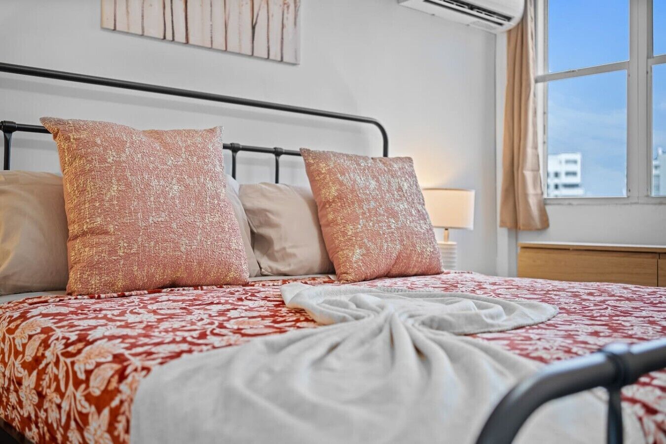 A hotel room with a bed, pillows, a nightstand, a lamp, and a window with a view of buildings.