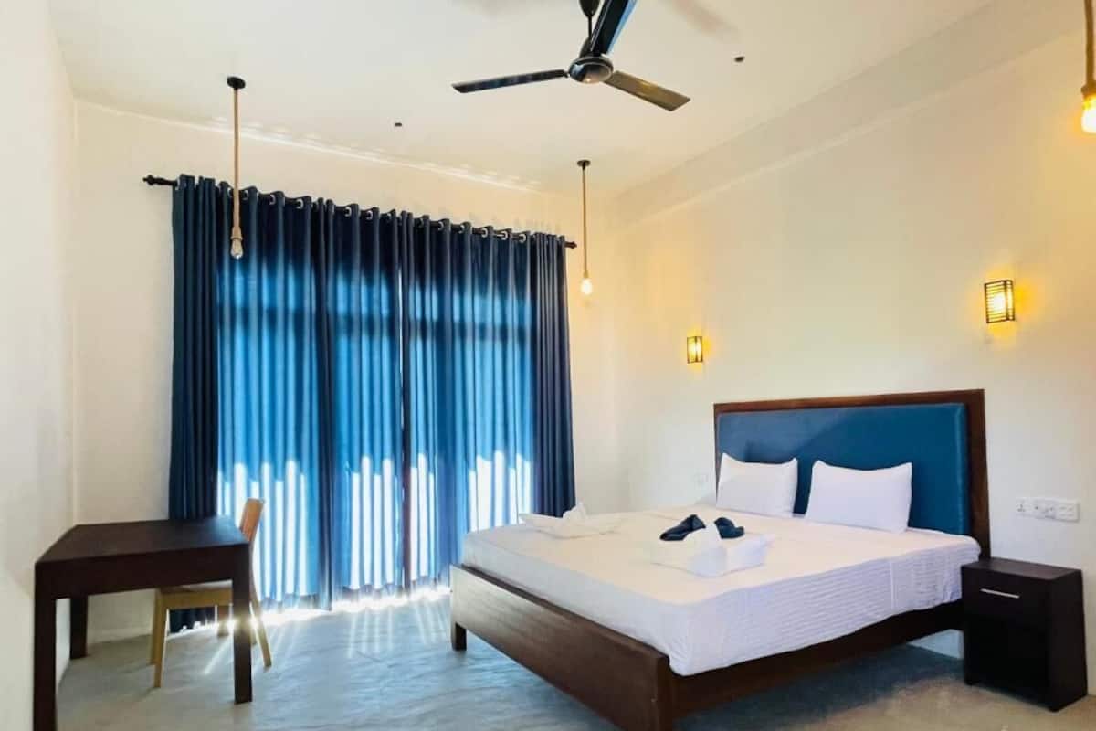 Habitación de hotel con cama, escritorio, silla, ventilador de techo y ventana con cortinas.