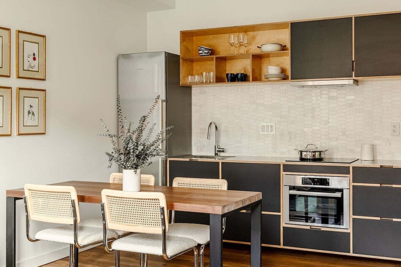 A modern kitchen with a wooden dining table, two wicker chairs, a built-in oven, and a stainless steel refrigerator.