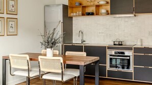 A modern kitchen with a wooden dining table, two wicker chairs, a built-in oven, and a stainless steel refrigerator.