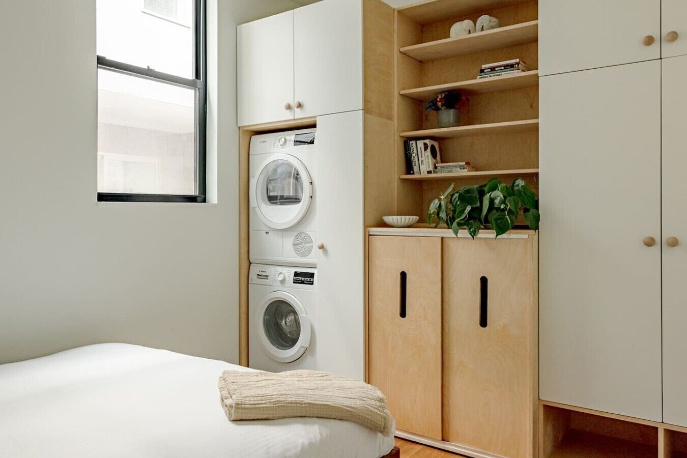 A bedroom with a bed, a washing machine, wooden cabinets, and a window.