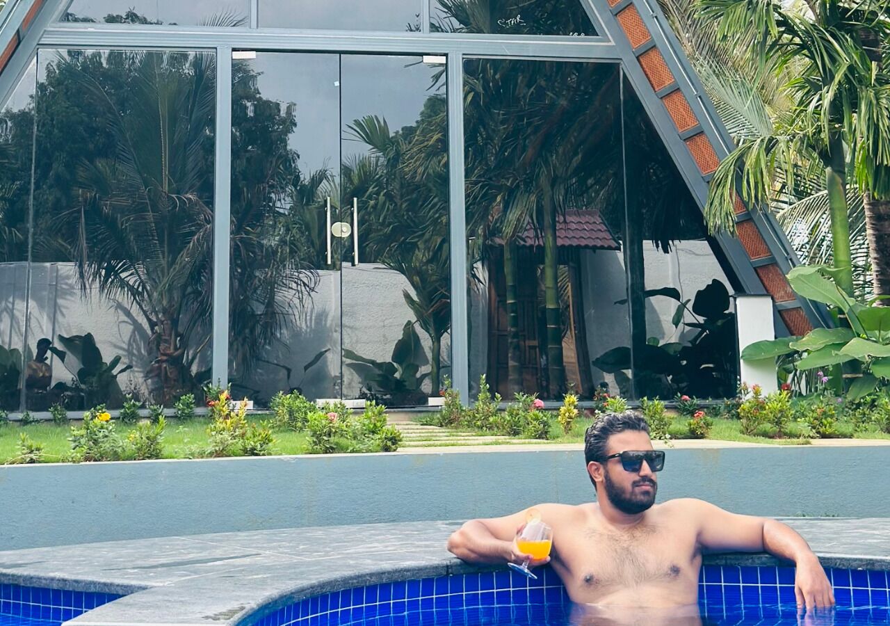 Un homme se détend au bord d’une piscine, un verre à la main, entouré de verdure et d’un bâtiment moderne aux fenêtres en verre.