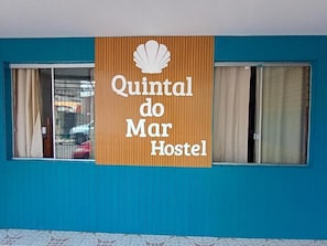 Placa de hostel com um logotipo de concha e o nome 'Quintal do Mar Hostel' em uma parede azul.