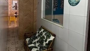A narrow hallway with patterned floor tiles, a wooden bench with a leaf-patterned cover, a window with a view of a building, and a trash bin. - Quintal do Mar Hostel (Salvador)