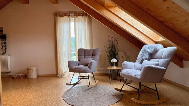 Une pièce mansardée chaleureuse avec des poutres en bois, deux fauteuils à bascule, une petite table avec une lampe et une vue sur l’extérieur par une fenêtre ornée de rideaux.