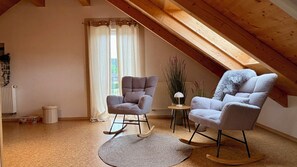 Ett mysigt rum på vinden med synliga träbjälkar, två gungstolar, ett litet bord med en lampa och en utsikt över omgivningarna genom ett fönster med gardiner.