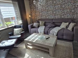 A living room with a sofa, armchair, coffee table, and a stone wall. - Eifelglück House  (Großlittgen)