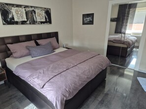 A modern bedroom with a large bed, a nightstand, a bedside table, a wall-mounted artwork, and a framed picture. - Eifelglück House  (Großlittgen)