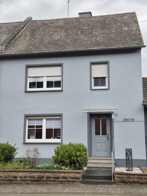 A two-story building with a grey facade, white-framed windows, a grey door, and a small garden area with bushes. - Eifelglück House  (Großlittgen)