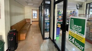 A hallway with a brown sofa, a black luggage, and a sign indicating a 24-hour mart. - Cozzy Galaxy Hotel (Manado)