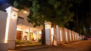 Un hotel moderno con una entrada bien iluminada, un espacio exterior cubierto con una hamaca y un árbol grande que da sombra.