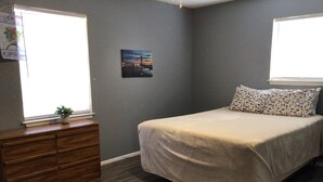 Un dormitorio con cama, cómoda, ventanas y un cuadro colgado en la pared.