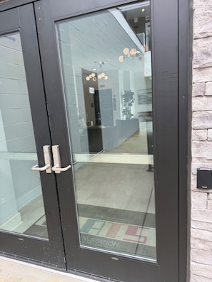 A black-framed glass door with a handle, reflecting a hallway with a patterned floor and a plant.