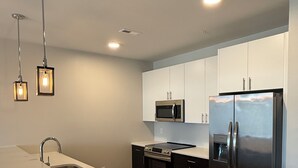 A modern kitchen with a stainless steel refrigerator, a microwave, and a dishwasher. There are pendant lights above the sink and a large island with a sink. - NEW Designer Coastal Condo - Sleeps 8 -Close to Downtown Rehoboth Next to Dining (Rehoboth Beach)
