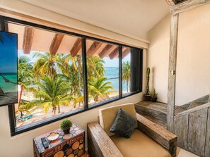 A room with a large window overlooking a beach, a wooden bench, a sofa with cushions, and a small table with a plant and a remote control.