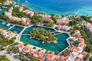 Vista aérea de un complejo turístico frente al mar, con varias edificaciones, un puerto deportivo y aguas azules cristalinas.