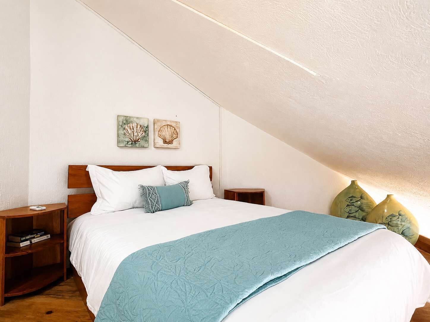 A bedroom with a sloped ceiling, a bed with white bedding and a teal throw, wooden nightstands, and two wall-mounted paintings.