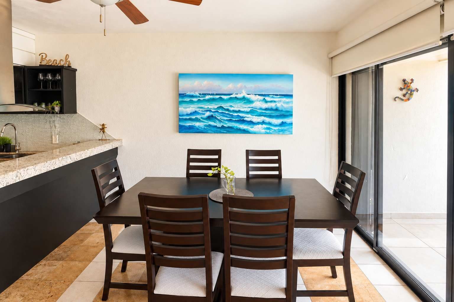 Zona pranzo con tavolo e sedie, un quadro raffigurante l'oceano, angolo cottura con piano di lavoro e pensili, e un elemento decorativo a parete.