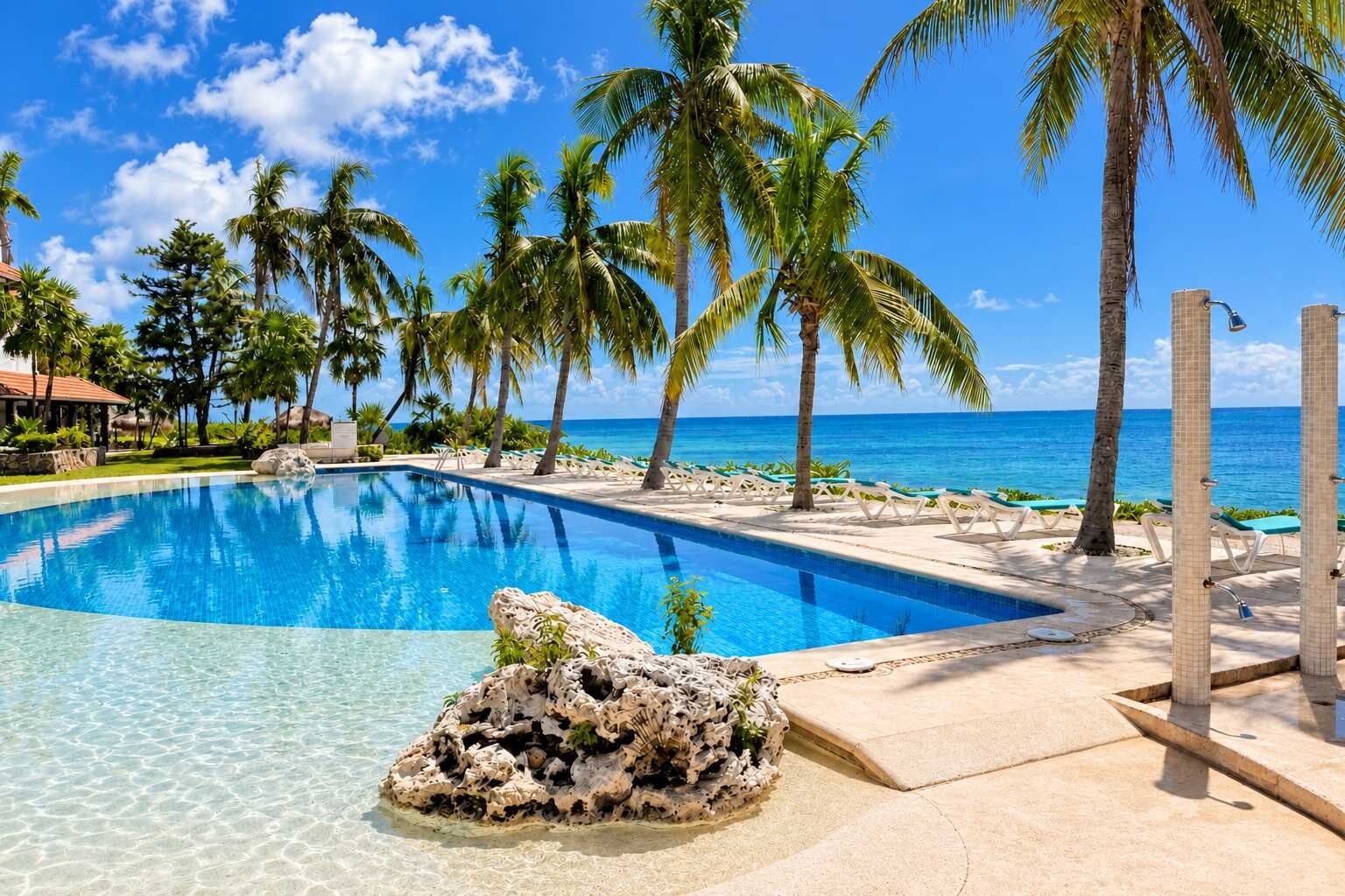A clear blue pool with a rock formation in the center, surrounded by palm trees and a view of the ocean.
