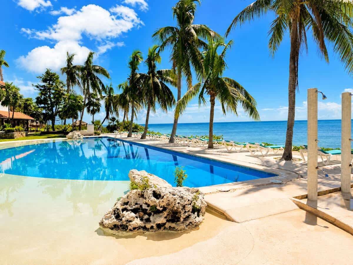 Una piscina azul cristalina rodeada de palmeras y una playa, con una formación rocosa al frente.
