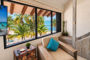 Une pièce dotée d’une grande fenêtre donnant sur une plage, d’un canapé avec un coussin, d’une petite table avec une plante et d’un coffre ancien.
