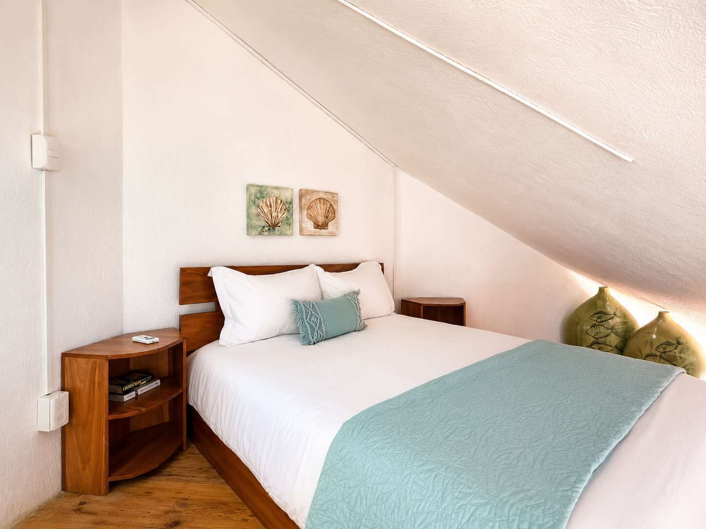 Une chambre avec un plafond en pente, une tête de lit en bois, une literie blanche, un plaid turquoise, deux œuvres d’art accrochées au mur et une table de chevet avec un livre.