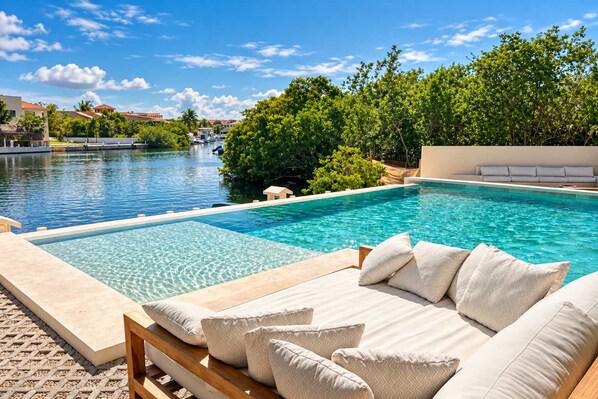 A swimming pool with a wooden deck and lounge area, surrounded by greenery and a clear sky. - Canal view, pool, 7-min walk to beach (Puerto Aventuras)